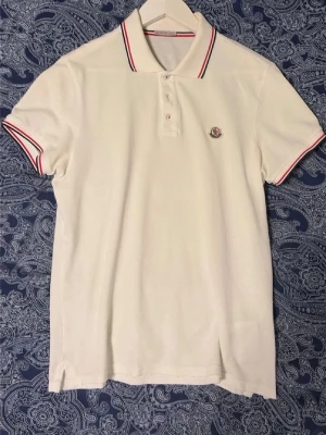 Moncler pike - Vit pikétröja från Moncler med klassisk krage och knäppning framtill. Tröjan har röda och blå ränder på krage och ärmslut samt Moncler-logga på bröstet. Den är äkta och är i fint skick hör av vid fler frågor