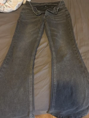 Svarta bootcut jeans med vit söm - Säljer ett par svarta bootcut jeans med tydliga vita sömmar och låg midja. Jeansen har klassisk femficksdesign och dragkedja med knapp. Perfekta för dig som gillar en retro eller alternativ stil.