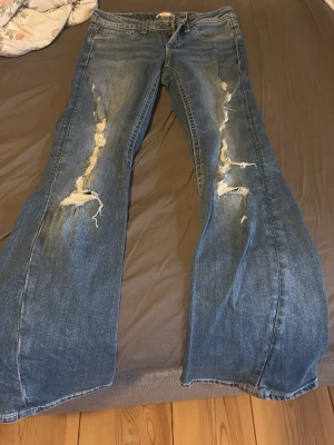 Blå slitna bootcut jeans - Säljer ett par blå bootcut jeans med slitningar och hål längs båda benen. Jeansen har klassisk femficksdesign, knappgylf och är tillverkade i denim. Perfekta för en avslappnad och trendig look.