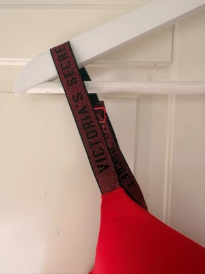 Röd push-up bh från Victoria's Secret - Snygg röd push-up bh från Victoria's Secret med vadderade kupor och svarta axelband med logotyp. Bh:n har en modern design och ger bra stöd. Perfekt för dig som vill ha en färgstark och bekväm bh med sportig känsla.