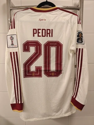 Spanien spelar Version landslagströja Pedri #20 - Vit långärmad Spanien spelar Version landslagströja från Adidas med Pedri och nummer 20 på ryggen. Tröjan har röda detaljer, guldinslag vid krage och ärmslut samt officiella FIFA och turneringsmärken på ärmarna. Perfekt för fotbollsfans som vill sticka ut på läktaren.