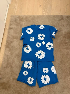 Blått set från denim tears - Snyggt blått set med stora vita blommiga mönster från denim tears. Setet består av en t-shirt med rund hals och korta ärmar samt matchande shorts med resår i midjan. Perfekt för en avslappnad och trendig look hemma. L/M