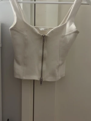 Vit zara topp - Snygg vit topp från zara med markerade sömmar och en rejäl dragkedja framtill. Toppen är smått croppad och har breda axelband, perfekt att styla med höga jeans eller kjol.