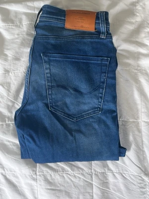 Blå jeans från Jack & Jones - Snygga blå jeans från Jack & Jones med klassisk femficksdesign och läderpatch bak i midjan. Jeansen har en rak passform och är tillverkade i stretchigt denimtyg för extra komfort. Perfekta till en avslappnad stil.