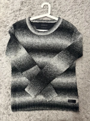 Gråmelerad stickad tröja Tiger of Sweden - Säljer en stickad tröja från Tiger of Sweden Jeans i storlek S. Tröjan har breda horisontella ränder i olika nyanser av grått och svart, rund halsringning och ribbade muddar. Perfekt för dig som gillar en stilren och mysig look. Har ett litet hål vid nacken som knappt syns annars inga andra defekter 