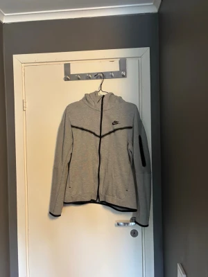Grå nike tech fleece - Snygg grå hoodie från Nike med svart dragkedja och svarta detaljer. Hoodien har en huva, lång ärm och en diskret Nike-logga på bröstet. Materialet är mjukt och stretchigt, perfekt för en sportig och avslappnad stil.