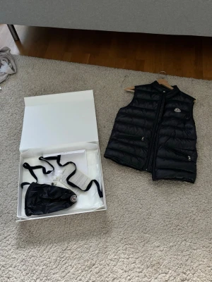 Svart guiväst från Moncler - Snygg svart dunväst från Moncler med klassisk quiltad design och hög krage. Västen har två dragkedjeförsedda fickor och Moncler-logga på bröstet. Kommer med originalpåse och box. Perfekt för lager-på-lager-stil under kyliga dagar.