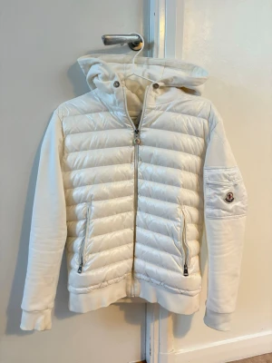 Vit pufferjacka med huva från Moncler - Snygg vit pufferjacka från Moncler med quiltad front och huva. Jackan har dragkedja framtill, två fickor med dragkedja och Moncler-logga på ärmen. Saknar snöre.