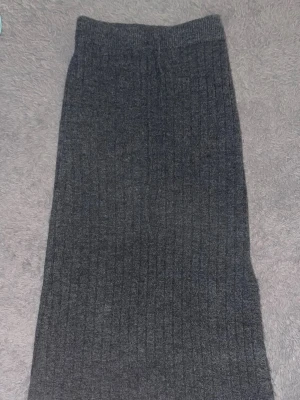 Grått ribbstickat set med tröja & kjol - Mysigt tvådelat set i grå ribbstickad design. Setet består av en långärmad tröja med polokrage och en matchande lång kjol. Perfekt för dig som gillar en enkel och stilren look. Materialet är mjukt och stretchigt för extra komfort.