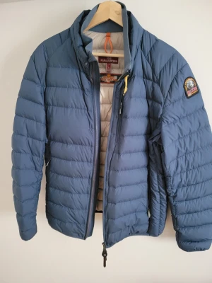 Blå dunjacka från Parajumpers - Snygg blå dunjacka från Parajumpers med quiltad design och dragkedja framtill. Jackan har en klassisk ståkrage, orange detalj i nacken och logotyp-patch på ärmen. Perfekt för kalla dagar och riktigt skön att bära.
