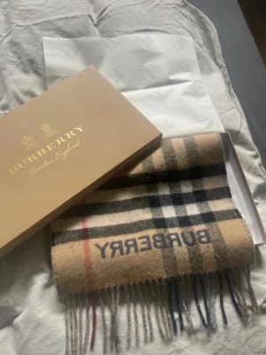 Burberry halsdukar i ull - Klassisk halsduk från Burberry i mjuk ull. Halsduken är beige med ikoniskt rutmönster och svart BURBERRY-logga, och mörkgrå med rutmönster och ljus logga. Fransar i ändarna och levereras i originalkartong.