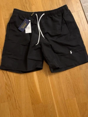 Svarta badshorts från Polo Ralph Lauren - Svarta badshorts från Polo Ralph Lauren med vit snörning i midjan och klassisk logga broderad på ena benet. Shortsen har elastisk midja och sidofickor, perfekta för en avslappnad stil. Pris kan diskuteras vid snabb affär. 
