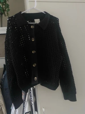 Svart stickad kofta från H&M - Svart stickad kofta från H&M med stora knappar framtill och klassisk krage. Koftan har ett genomgående hålmönster och ribbade muddar vid ärmslut och nederkant. 