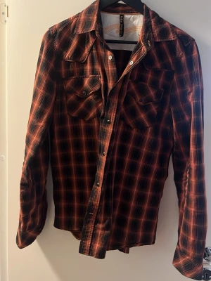 Rutig skjorta från Nudie Jeans - Snygg långärmad skjorta från Nudie Jeans i rött och svart rutigt mönster. Skjortan har två bröstfickor med knappar och klassisk krage. Perfekt för en avslappnad och cool stil.