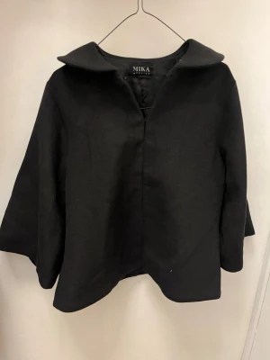 Svart kappa från MIKA Atelier - Stilren svart kappa från MIKA Atelier med bred krage och trekvartsärmar. Kappan har en minimalistisk design utan synliga knappar och är tillverkad i ett mjukt, tjockt tyg som passar perfekt för kyligare dagar. Passar xs-M. Nypris 1600