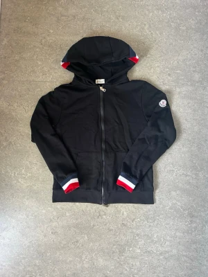 Svart Moncler hoodie med dragkedja - Svart hoodie från Moncler med hel dragkedja, huva och fickor framtill. Ärmsluten och insidan av huvan har snygga detaljer i rött, vitt och blått. Moncler-logga på ena ärmen. Perfekt för en stilren och sportig look.