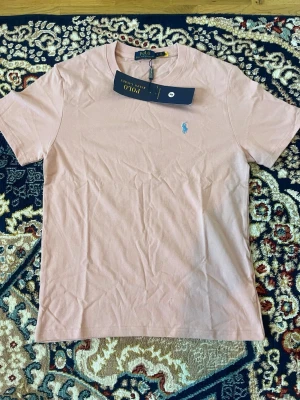 Ljusrosa t-shirt från Polo Ralph Lauren - Snygg ljusrosa t-shirt från Polo Ralph Lauren med klassisk passform och broderad blå logga på bröstet. Tillverkad i mjuk bomull och har rund halsringning samt korta ärmar. Perfekt för en clean och stilren look.