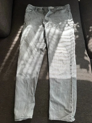 Ljusgrå raka jeans - Säljer ett par ljusgrå jeans med rak passform och klassisk femficksdesign. Jeansen har bälteshällor och knappgylf. Perfekta för en avslappnad och stilren look. Materialet är denim i bomull. Modell= Relaxed/ Chris och är storlek 29/34. Har ett hål en bit nedanför under knapparna, lätt att sy ihop.