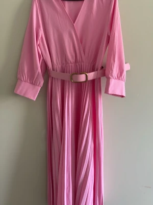 Lång rosa omlottklänning med bälte - Snygg lång rosa klänning med omlottliv och v-ringning. Klänningen har trekvartsärmar, plisserad kjol och matchande bälte i midjan med guldfärgat spänne. Perfekt för dig som gillar en feminin och trendig stil.