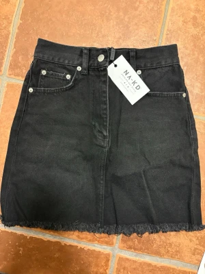 Svart jeanskjol från NA-KD - Trendig svart jeanskjol från NA-KD med rå kant nertill och klassisk femficksdesign. Kjolen har knapp- och dragkedjestängning framtill och är perfekt för dig som gillar en enkel men snygg look. Oanvänd med lappen kvar. Storlek XXS. 