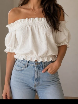 Vit offshoulder blus med volang - Superfin vit offshoulder blus med puffärmar och volangdetaljer vid halsringning och nederkant. Blusen har en smockad resår upptill och nedtill som ger en snygg passform. Perfekt till jeans eller kjol för en trendig look.