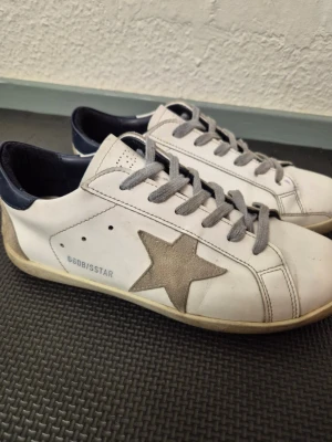 Golden Goose vita sneakers med stjärna. Pris kan diskuteras - Golden Goose sneakers i vitt läder med grå mockadetaljer och ikonisk stjärna på sidan. Skorna har grå skosnören, mörkblå hälkappa och låg profil. Snygg kontrast mellan material och färger, perfekt för dig som gillar streetstyle och unika detaljer.