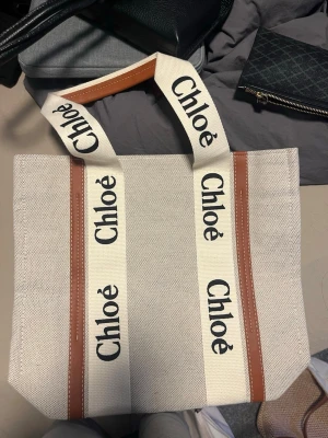 Chloé beige väska  - Trendig shoppingväska från Chloé i beige canvas med breda vita axelband prydda med svart Chloé-logga. Väskan har bruna läderdetaljer och rymligt innerfack med ljus textilfodring. Perfekt för dig som vill ha en stilren och lyxig tote att bära till vardags.