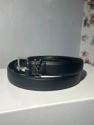 Svart YSL bälte i skinn - Stilrent svart bälte från Yves Saint Laurent i äkta skinn med silverfärgat YSL-spänne. Perfekt accessoar för att lyfta din outfit med en lyxig touch. Klassisk design med fyrkantigt spänne och diskreta sömmar.