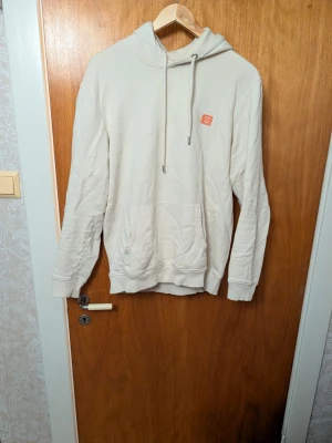 Beige hoodie från Les Deux - Snygg beige hoodie från Les Deux med huva och dragsko. Klassisk känguruficka framtill och liten orange logga på bröstet. Perfekt för en chill och stilren look. Mjuk bomullskänsla och relaxed fit.