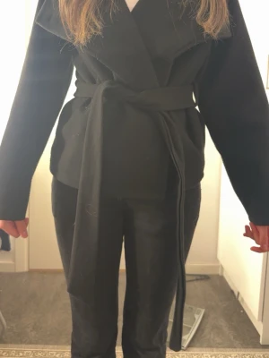 Svart kappa med knytskärp - Stilren svart kappa med bred krage och knytskärp i midjan. Modellen är kortare och har raka ärmar. Perfekt för dig som gillar en enkel men trendig look. Materialet ser ut att vara mjukt och något tjockare, passar bra till höst och vinter.