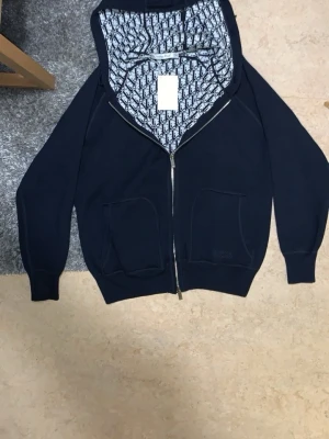L. Dior kofta/cardigan - Säljer denna kofta/cardigan för att har bytt ny stil till casual/Classic har inte plats för nya stilen i garderoben därför väljer och sälja Snygg marinblå hoodie från Christian Dior med dragkedja och två stora fickor framtill. Insidan av huvan har ett ikoniskt Dior-mönster i svart och vitt. Tillverkad i Italien och gjord i mjukt material som känns lyxigt och bekvämt. Perfekt för dig som vill ha något exklusivt och stilrent.