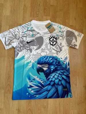 Nike Brasil CBF t-shirt med blå papegoja - Unik sporttröja från Nike och Brasilien CBF med färgstark blå papegoja och grafiskt mönster i vitt, grått och turkost. T-shirten har rund hals, kort ärm och är tillverkad i lätt, ventilerande Dri-FIT-material. Perfekt för dig som vill ha en snygg och skön fotbolls T-shirt. Den är helt ny och oanvänd, skriv vid frågor eller funderingar.