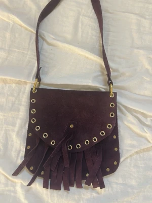 Lila axelväska i mocka med fransar - Cool liten lila axelväska i mocka med fransar och guldfärgade nitar runt locket. Väskan har en justerbar axelrem och stängs med ett lock. Perfekt för dig som gillar boho-stil och vill sticka ut med accessoarer.