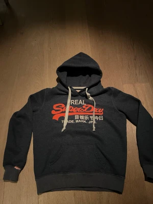 Mörkblå Superdry hoodie med tryck - Mörkblå hoodie från Superdry med stor orange logga och japansk text på bröstet. Klassisk känguruficka framtill och vita dragsnören i huvan. Mjuk bomullsblandning och ribbade muddar vid ärmslut och nederkant. Perfekt för en avslappnad streetwear-look.