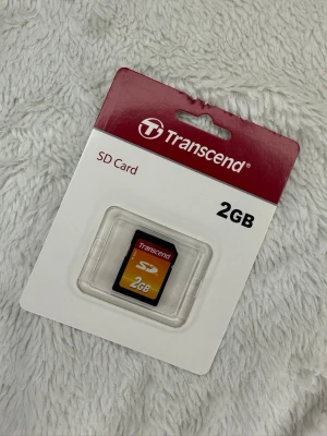 Transcend SD-kort 2GB - Transcend SD-kort med 2GB lagringsutrymme. Kortet är svart med röd och gul etikett och levereras i originalförpackning. Perfekt för kameror och annan elektronik som kräver SD-minneskort. Tillverkat i plast.