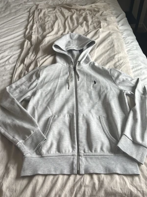 Ljusgrå hoodie från Polo Ralph Lauren - Snygg ljusgrå hoodie från Polo Ralph Lauren med dragkedja framtill och klassisk huva med dragsko. Broderad logga på bröstet och två fickor framtill. Tillverkad i mjukt bomullsmaterial som är skönt mot huden.