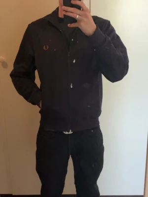 mörkblå ziptröja från Fred Perry - Snygg mörkblå ziptröja från Fred Perry med guldfärgad logga på bröstet. Tröjan har hög krage, ribbade muddar och dragkedja framtill. Perfekt till streetwear eller en avslappnad look.