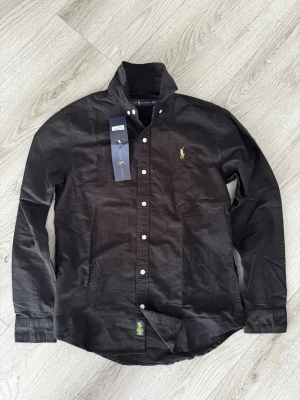 Svart skjorta från Ralph Lauren - Passar Storlek S.  svart skjorta från Ralph Lauren med vit knappstängning och ikonisk broderad logga på bröstet. Skjortan har lång ärm och normal passform, tillverkad i mjuk bomull. Perfekt för en stilren look.