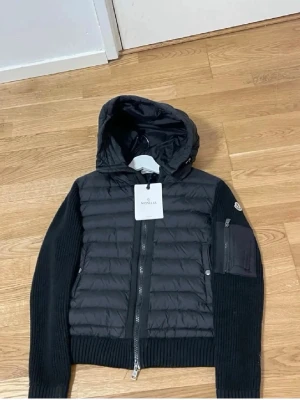 Svart pufferjacka från Moncler - Snygg svart pufferjacka från Moncler med stickade ärmar och huva. Jackan har dragkedja framtill, ribbade muddar och en praktisk ficka med dragkedja på ena ärmen. Perfekt för kalla dagar och riktigt trendig look.