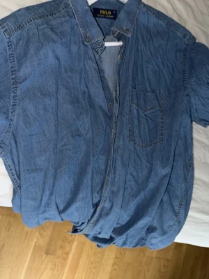 Blå skjorta från Polo Ralph Lauren - Säljer en klassisk blå skjorta från Polo Ralph Lauren i storlek XL. Skjortan har korta ärmar, knappkrage och bröstficka. Tillverkad i mjuk bomull med snygga sömdetaljer och knappar framtill. Perfekt för en avslappnad men stilren look.