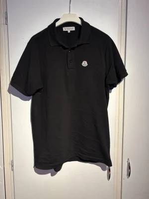 Svart pikétröja från Moncler (Äkta) - Klassisk svart pikétröja från Moncler med liten broderad logga på bröstet och knäppning vid halsen. Tröjan har korta ärmar och är tillverkad i bomull, perfekt för en stilren och avslappnad look.