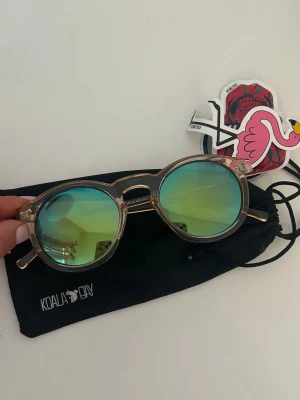 Runda solglasögon från Koala Bay - Runda solglasögon från Koala Bay med transparent beige båge och spegelglas i blågrön ton. Kommer med svart glasögonpåse och 3 stycken stickers. Köpta i Spanien för 24,95€. Aldrig använda. 