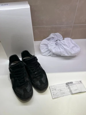 Svarta sneakers från Maison Margiela - Stilrena svarta sneakers från Maison Margiela med klassisk låg siluett. Skorna är tillverkade i en mix av mocka och läder, med diskreta detaljer och svart sula. Perfekta för dig som gillar minimalistisk och exklusiv design. Kan gå ner i pris vid snabb affär