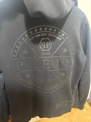 Svart hoodie från Philipp Plein - Snygg svart hoodie från Philipp Plein med dragkedja framtill och huva med dragsko. Stort ton-i-ton-tryck på ryggen med logga och text, . Tillverkad i mjukt bomullsmaterial och har fickor framtill. 