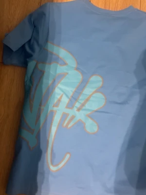Blå t-shirt från syna wrld - PASSAR L-XLSnygg blå t-shirt från Suckeraries med stort grafiskt tryck i ljusblått och beige på framsidan. Klassisk rund hals och korta ärmar. Perfekt för dig som gillar streetwear och vill sticka ut med en unik design.