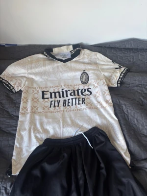 AC Milan bortatröja 2022/23 - Snygg AC Milan bortatröja från säsongen 2022/23 i vitt med beige mönster och svarta detaljer. Tröjan har klubbmärke och sponsortryck på bröstet samt klassisk krage. Perfekt för fotbollsträning eller att bära till vardags.