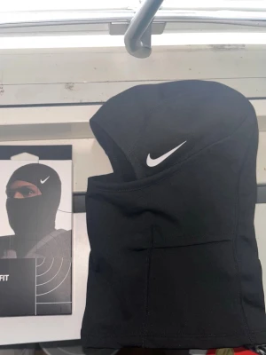 Nike Pro Therma-FIT balaclava - Svart balaclava från Nike med Therma-FIT-teknologi. Snygg och sportig design med vit Nike-logga framtill. Perfekt för träning utomhus eller kalla dagar. Materialet är mjukt och stretchigt för bästa komfort.