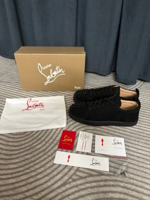 Svarta Christian Louboutin sneakers - Snygga svarta sneakers från Christian Louboutin med ikoniska röda sulor och svarta nitar på tån. Skorna är låga och har en lyxig känsla med ovandel i mocka och klassisk snörning. Perfekta för dig som vill sticka ut med exklusiv design.