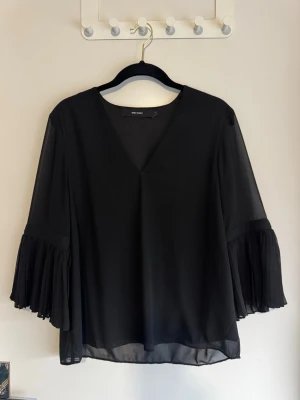 Svart blus med volangärm Vero Moda - Snygg svart blus från Vero Moda med v-ringning och vida, transparenta volangärmar. Blusen är tillverkad i ett lätt och luftigt material som ger en stilren och trendig look. Perfekt att matcha med jeans eller kjol för en cool outfit.