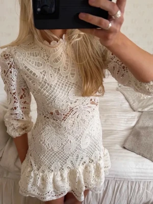 Vit spetsklänning med volang - Storlek S men passar xs.  Superfin vit kort klänning i spets med volangkant nertill och trekvartsärmar. Klänningen har hög hals och vackra broderade detaljer som ger en romantisk känsla. Perfekt för dig som vill sticka ut med en elegant look. Använd 0 gånger då jag blev sjuk när den skulle användas .
